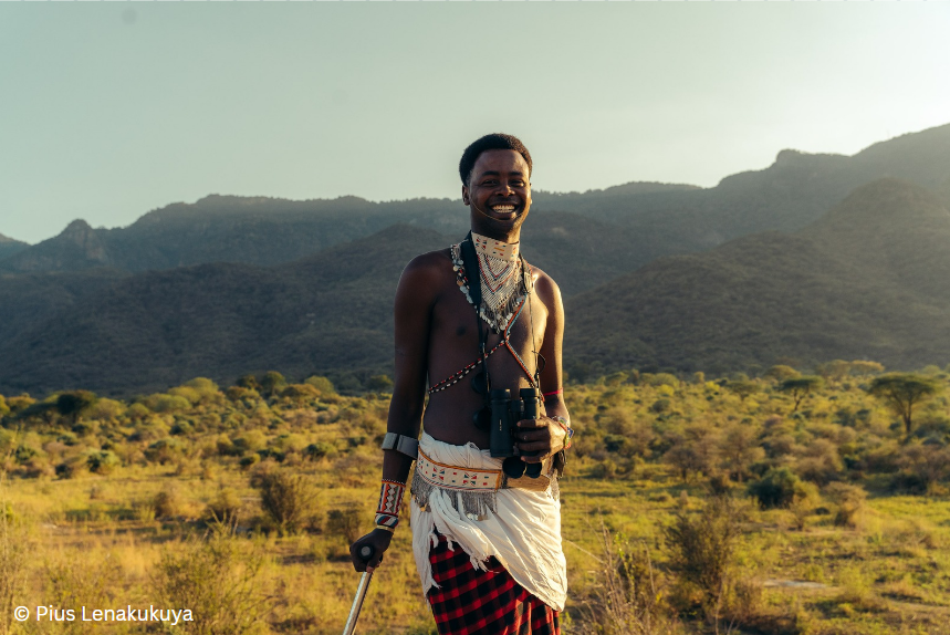 A man wearing traditional Kenyan dress is standing in a savanna, he is holding a walking stick. 